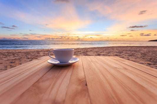 Kaffeetasse am Strand auf einem Tisch, Hintergrund Meer und Sonnenuntergang, Schriftzug "Die besten 5 Rezepte für Ihren perfekten Sommer mit Kaffee"