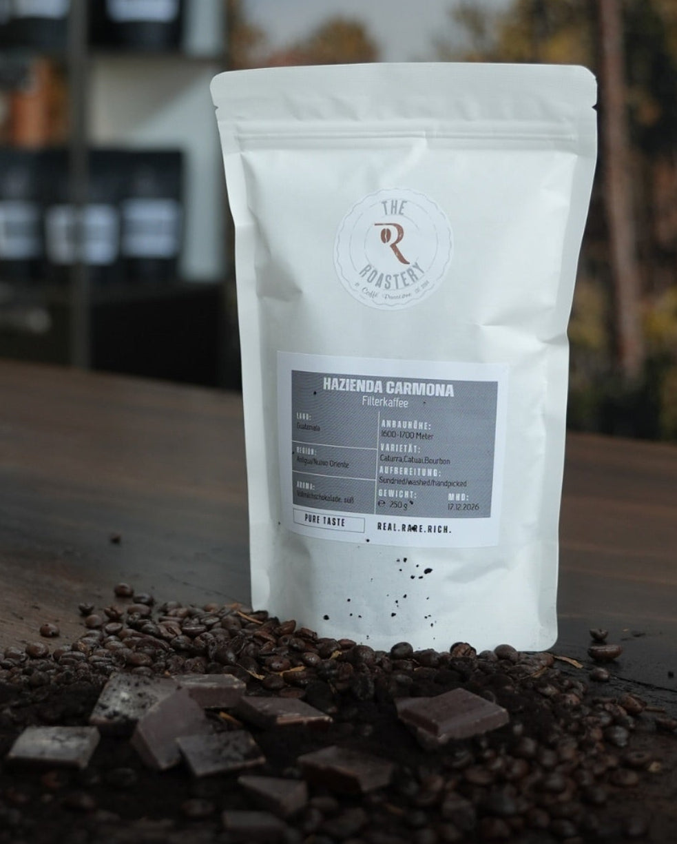 Weißer Filterkaffeebeutel „Hazienda Carmona“ auf rustikalem Holztisch, umgeben von gerösteten Kaffeebohnen und feiner Schokolade