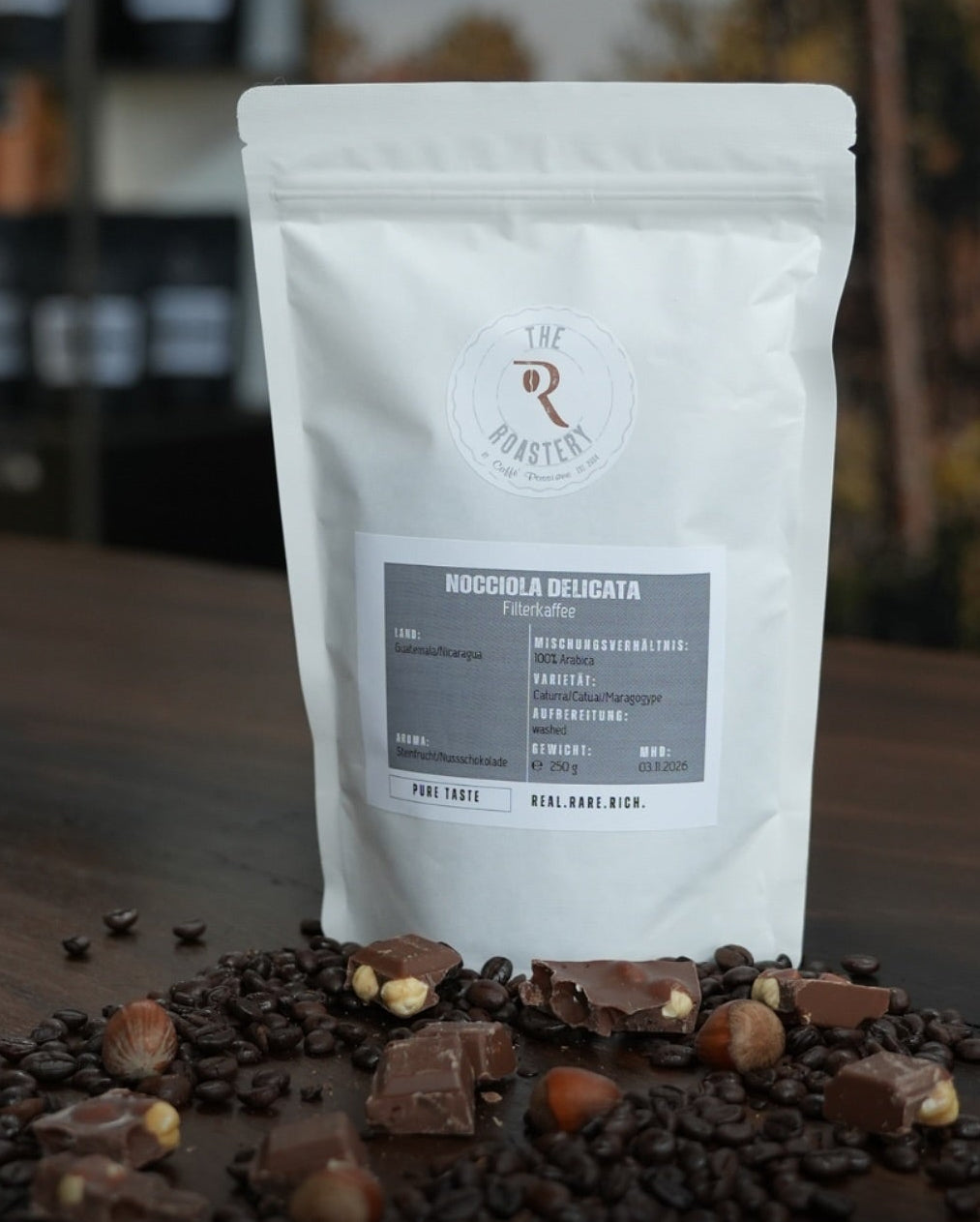 Weißer Filterkaffeebeutel „Nocciola Delicata“ auf rustikalem Holztisch, umgeben von gerösteten Kaffeebohnen und Nuss-Schokolade